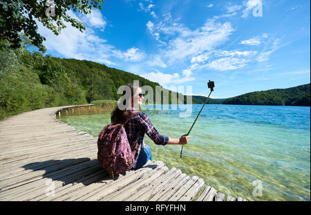 Colorful spring panorama of lit by bright sun green forest and clear blue lake. Young tourist woman in sunglasses and with backpack and camera sitting Stock Photo