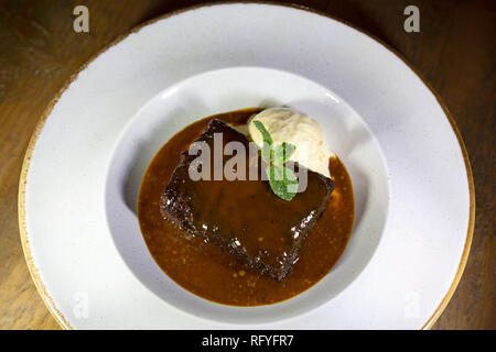 Sticky toffee pudding served with caramel sauce and a scoop of ice cream. The traditional British dessert is garnished with a sprig of mint. Stock Photo