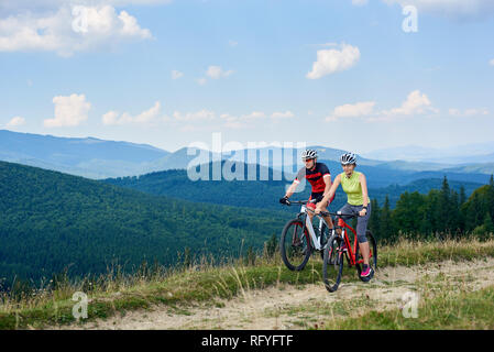 Young bikers tourists, man and woman in professional sportswear riding bikes down grassy field road under beautiful bright blue sky on magnificent mou Stock Photo