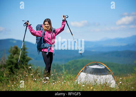 Happy woman tourist with backpack and trekking sticks near tent, wearing sports wear, standing on the top of hill against blue sky and clouds, rising  Stock Photo
