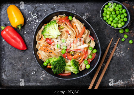 Vegetable stir fry with udon noodles, broccoli, carrot, pepper and green peas in bowl on black background. Top view Stock Photo