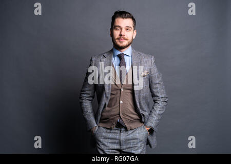 A confident elegant handsome young man standing in front of a grey background in a studio wearing a nice suit. Stock Photo