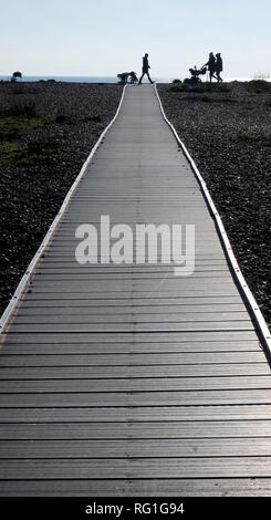Three silhouetted unrecognisable people and a baby in a pram walking across the end of a long straight wooden pathway with a pebble beach on both side Stock Photo