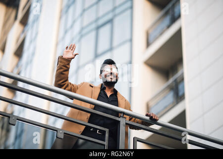 Serious indian businessman in eye glasses looking on copy space while standing against glass skyscraper waving hello to someone. Stock Photo