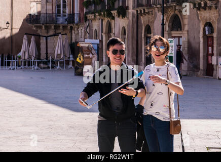Portrait of male and female tourists in town square, man holding selfie stick, laughing, Ortygia, Syracuse, Sicily, Italy, Europe Stock Photo