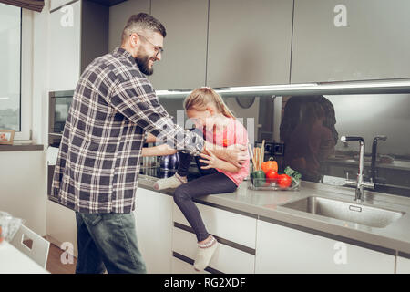 Cute long-haired girl in a pink t-shirt having fun with her father in the kitchen Stock Photo
