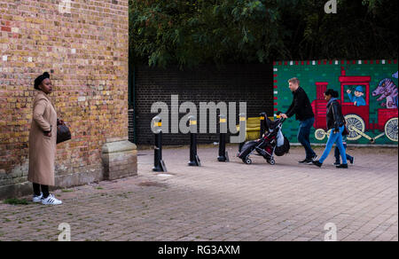 Woman standing beside brick wall, looking into distance, young family walking in background, pushing stroller, Crystal Palace, London, England, UK Stock Photo