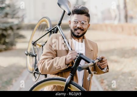 Bearded indian hipster man with a fixie bicycle. Low angle view of confident young bearded man carrying his bicycle on shoulder and looking at camera  Stock Photo