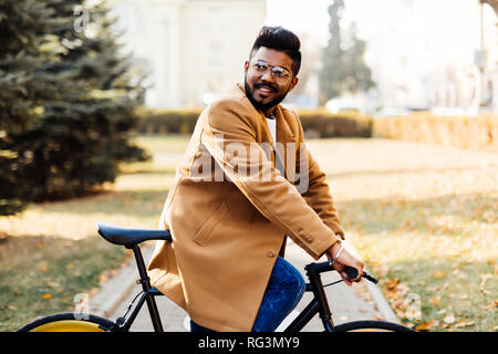 Young bearded indian man in coat while sitting on his bicycle outdoors Stock Photo