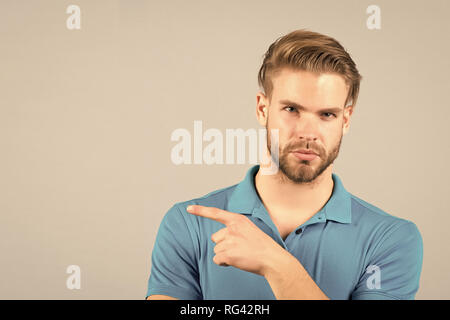 Advice advertisement concept. Guy bearded attractive pointing index finger to side. Man strict face looks confidently, grey background. Man with beard unshaven guy looks handsome well groomed. Stock Photo