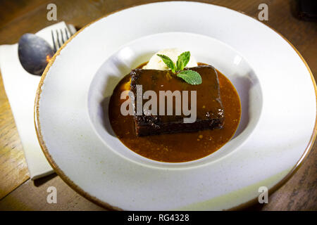 Sticky toffee pudding served with caramel sauce and a scoop of ice cream. The traditional British dessert is garnished with a sprig of mint. Stock Photo