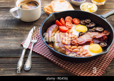 Fried eggs, sausages, bacon, beans and mushrooms in iron skillet, toasts, coffee, butter and jam on rustic wooden background. Full english breakfast. 