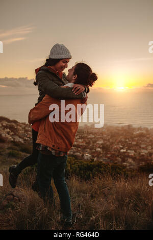 Romantic couple standing on mountain peak at sunset. Man carrying his girlfriend in his arms on cliff. Stock Photo