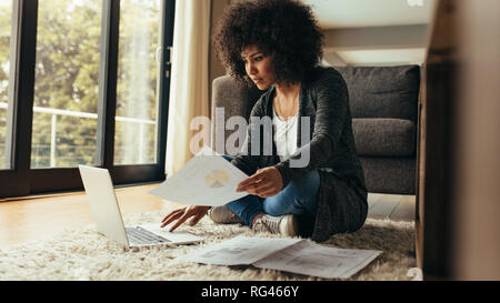African woman sitting on floor at home with document in hand using laptop. Woman with curly hair working from home. Stock Photo