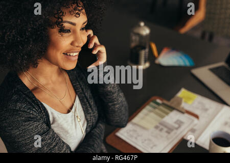 Close up portrait of happy young woman sitting at her desk talking on mobile phone. Female architect talking with client over smart phone at home offi Stock Photo