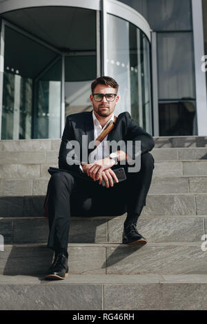 Entrepreneur sitting on the steps of a building holding his mobile phone. Businessman sitting on steps outdoors while travelling to office. Stock Photo