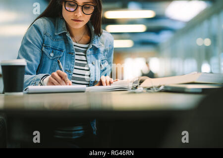 Woman student writing down notes in notepad from a textbook. University student studying at college library. Stock Photo