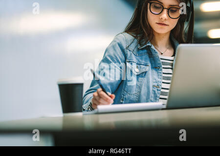 University student writing in a book while sitting at desk with laptop and coffee up at college campus. Female student studying at college library. Stock Photo