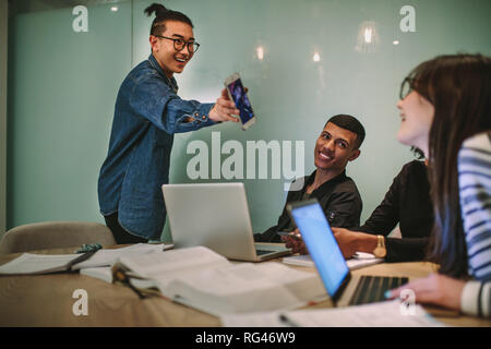 Man showing his mobile phone to female classmate while studying in college. Group of students at college campus looking at smartphone and smiling. Stock Photo