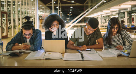 Multi-ethnic students sitting at college library. Students using making notes, using laptop and mobile phones while studying together. Stock Photo