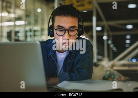 Asian male student with headphones sitting at library reading book and using laptop. Man studying at university library. Stock Photo