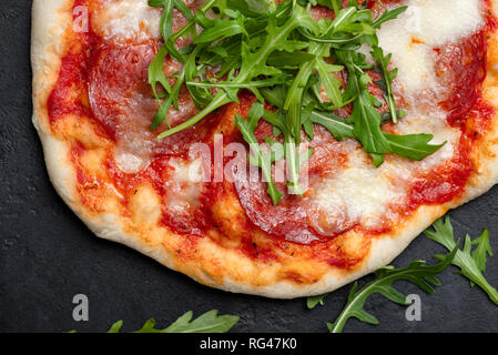 Pizza with arugula, salami sausage, mozzarella cheese and tomato sauce on black concrete background. Closeup view Stock Photo