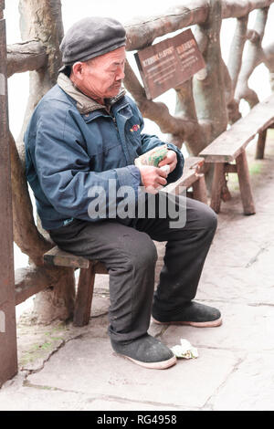 FENGHUANG,HUNAN,CHINA -DECEMBER 12 2018 : Unidentified old senior sitting on a bench counting his money on the bank at Tuojiang River in Fenghuang Stock Photo