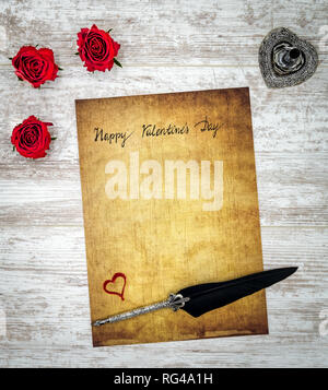 Classic Happy Valentine’s Day card hand written on parchment 3 red roses red painted hart ornate silver quill and stand white painted oak top view Stock Photo