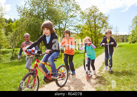 Children do a race with scooter and bike in the park in summer vacations Stock Photo