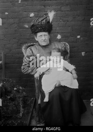 Older Edwardian lady sitting in a wicker chair in a backyard with a small baby on her knee, circa 1910. Stock Photo