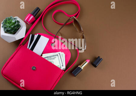 top view contents of women  bag  layout nail polish,lipstick, money,credit cards   and sunglasses in an open red leather  bag on brown background top  Stock Photo