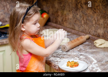 Little beautiful cute girl in orange apron smiling and making homemade pizza, roll out the dough at home kitchen. Concept happy family holiday, Italia Stock Photo