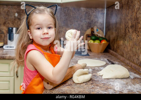 Little beautiful cute girl in orange apron smiling and making homemade pizza, roll out the dough at home kitchen. Concept happy family holiday, Italia Stock Photo