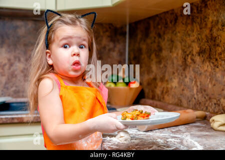 Little beautiful cute girl in orange apron smile and keep homemade pizza, roll out the dough at home kitchen. Concept happy family holiday, make and c Stock Photo