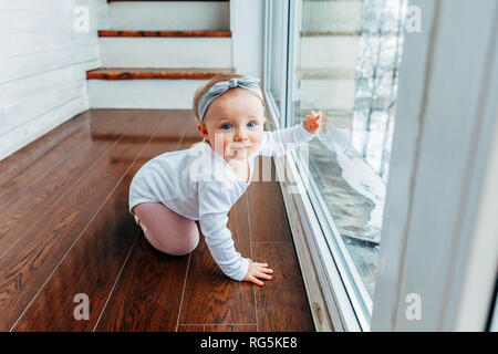 Little crawling baby girl one year old siting on floor in bright light living room near window smiling and laughing. Happy toddler kid playing at home Stock Photo