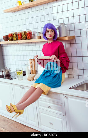 housewife with purple hair sitting on kitchen counter with rolling pin Stock Photo