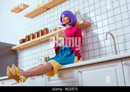 selective focus of housewife with purple hair and colorful clothes sitting on kitchen counter and holding glass jar of lemonade Stock Photo