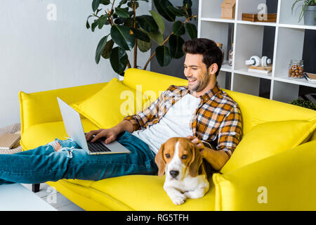 handsome laughing man using laptop on sofa with beagle dog Stock Photo
