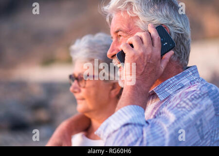 Happy retired couple of caucasian man and woman people hug and look in front of them using and speaking with modern smart technology phone - modern li Stock Photo