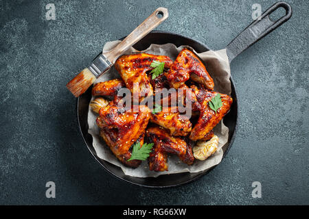 Baked chicken wings in barbecue sauce with sesame seeds and parsley in a cast iron pan on a dark concrete table. Top view Stock Photo