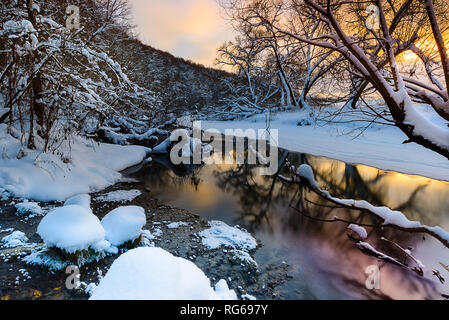 The river stretches into the distance, it reflects the sunset sky and trees, in the foreground stones covered with snow Stock Photo