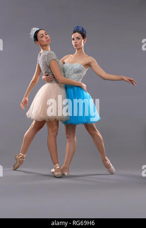 Two young ballerinas standing on socks, hugging each other and looking at camera in studio. Professional ballet dancers posing on grey isolated background. Concept of ballet dancing. Stock Photo