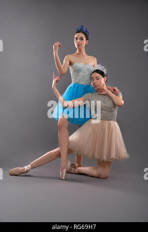 Two young graceful ballet dancers in dresses and ballet shoes performing on grey isolated background. Ballerina looking at camera and standing near partner sitting on floor. Concept of ballet.