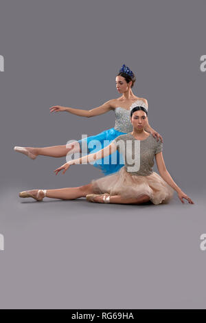 Front view of serious ballerina in beige dress sitting on floor and raising hand aside while female partner posing behind in studio. Professional dancers performing together. Concept of elegance.