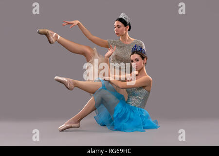 Two beautiful ballerinas sitting on foor in studio and posing. Graceful dances raising one leg and looking at camera on grey isolated background. Concept of classical art and ballet. Stock Photo