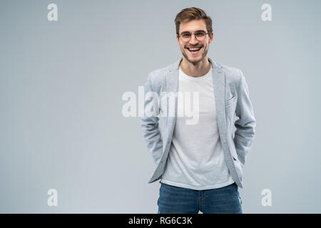 Portrait of attractive masculine fashionable modern stylish guy wearing blue blazer, jeans keeping hands in pockets isolated on gray background. Stock Photo