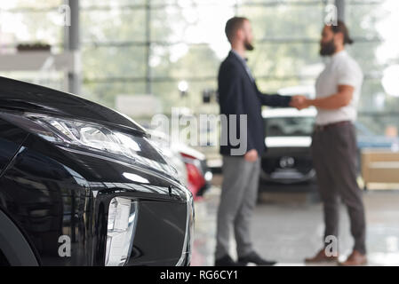Unrecognizable men shaking hands, standing in modern car showroom. Black car headlight. Car dealer and client of car center making deal. Client purchasing new vehicle.   Stock Photo