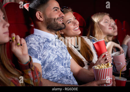 Close up of brunette Arabian man in printed blue shirt and blonde smiling woman looking at screen in cinema, Couple of positive friends having fun, spending time together. Concept of enjoyment, Stock Photo