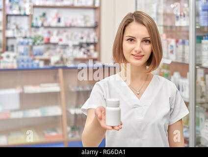 Beautiful woman posing in chemists store. Female pharmacist wearing in white uniform holding white plastic bottle for pills and medicines, showing it. Pretty woman looking at camera and smiling. Stock Photo