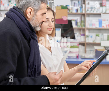 Male customer looking at black folder, listening to pharmacist. Woman in white uniform helping to clients of chemists shop. Mature bearded man looking for medicines. Stock Photo